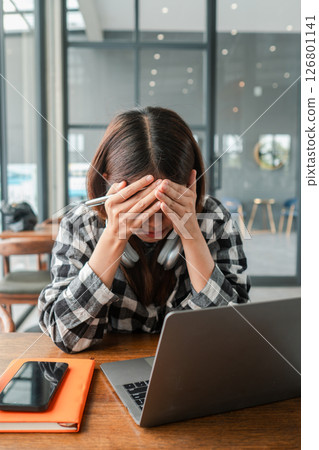 Young woman in checkered shirt feeling stressed while working on a laptop in a modern office setting. 126801141