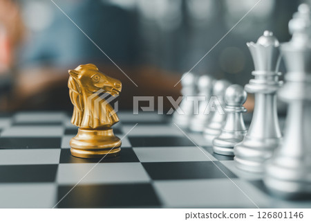 Close-up of a golden knight chess piece on a chessboard, surrounded by silver pieces, highlighting strategy and competition. Close-up of a golden knight chess piece on a chessboard, surrounded by silver pieces, highlighting strategy and competition. 126801146