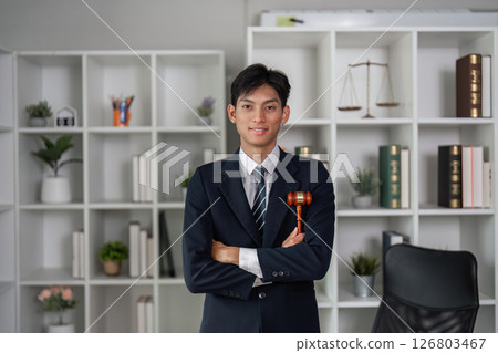Confident young Asian lawyer standing with arms crossed in office, holding gavel, representing law practice. 126803467