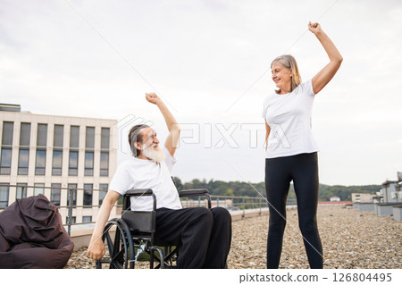Senior man in wheelchair and woman performing fitness stretches together on outdoor rooftop. Smiling expressions suggest positivity, unity. Concept showcases active lifestyle, aging gracefully 126804495