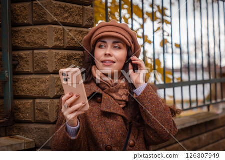 Portrait of Caucasian smiling woman in coat and hat using smartphone and looking at the left side. Road in background. Autumn city modern lifestyle Portrait of Caucasian smiling woman in coat and hat using smartphone and looking at the left side. Road in background. Autumn city modern lifestyle 126807949