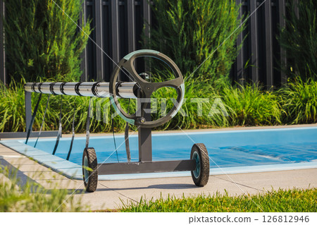 Manual pool cover reel system on wheels next to a private outdoor swimming pool, with green landscaping in the background 126812946