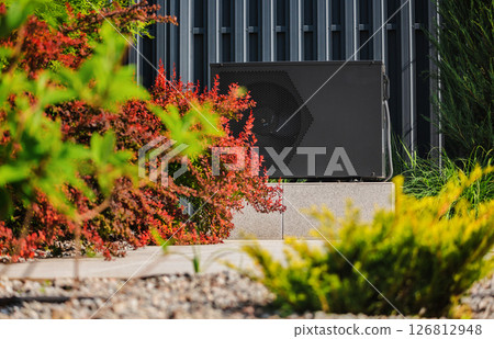 Outdoor heat pump unit placed on a concrete base in a landscaped garden, with colorful bushes and modern metal wall in the background 126812948