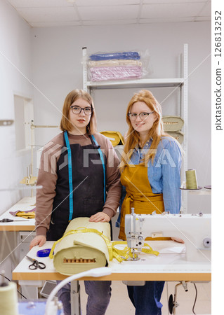 Two female seamstresses in workshop at factory are standing at a table with sewing machines.  126813252