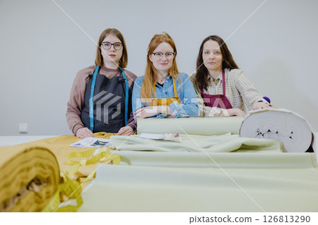 Three female seamstresses and fashion designers are standing at a table with a roll of fabric 126813290