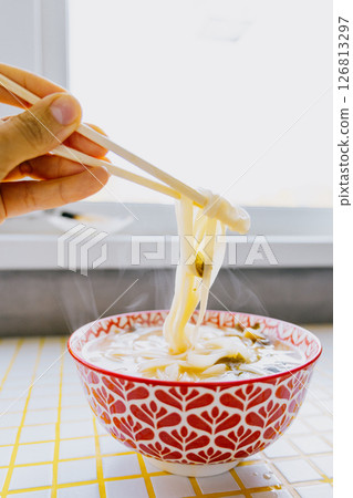 person is eating noodles with chopsticks. colorful bright red plate of ramen sits on yellow table. person is eating noodles with chopsticks. colorful bright red plate of ramen sits on yellow table. 126813297
