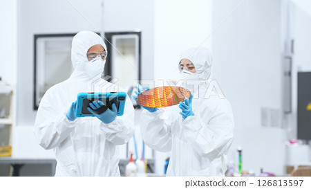 Male and female workers in protective clothing holding tablets and inspecting silicon wafers in a semiconductor factory Male and female workers in protective clothing holding tablets and inspecting silicon wafers in a semiconductor factory 126813597