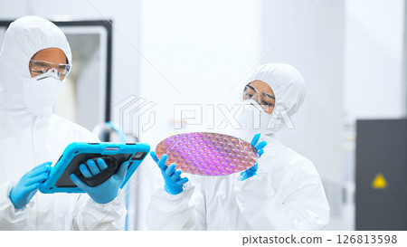 Male and female workers in protective clothing holding tablets and inspecting silicon wafers in a semiconductor factory Male and female workers in protective clothing holding tablets and inspecting silicon wafers in a semiconductor factory 126813598