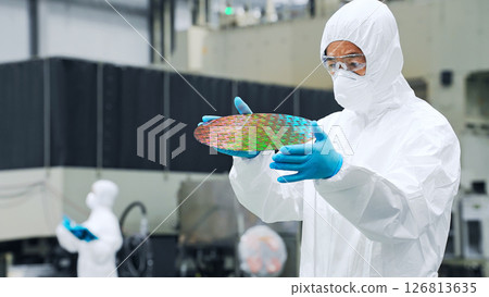 A male worker in a dustproof suit inspecting silicon wafers in a semiconductor factory A male worker in a dustproof suit inspecting silicon wafers in a semiconductor factory 126813635