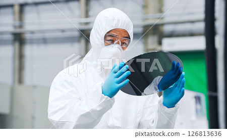 A male worker in a dustproof suit inspecting silicon wafers in a semiconductor factory A male worker in a dustproof suit inspecting silicon wafers in a semiconductor factory 126813636