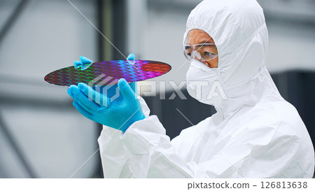 A male worker in a dustproof suit inspecting silicon wafers in a semiconductor factory A male worker in a dustproof suit inspecting silicon wafers in a semiconductor factory 126813638