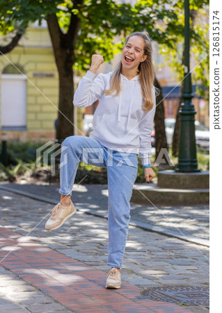 Young woman looking surprised clenching fists, shocked by sudden victory standing on city street 126815174