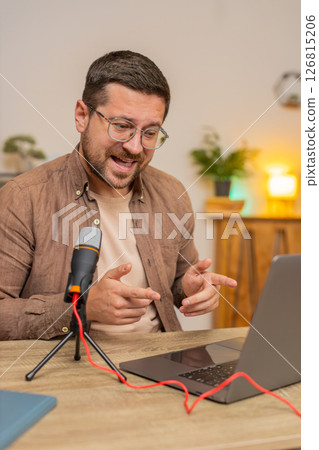 Happy young Caucasian man influencer speaking into microphone and laptop webcam at home office table 126815206