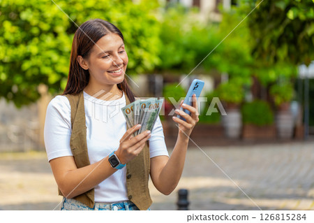 Happy Caucasian woman counting money dollar cash, using smartphone calculator app on city street Happy Caucasian woman counting money dollar cash, using smartphone calculator app on city street 126815284