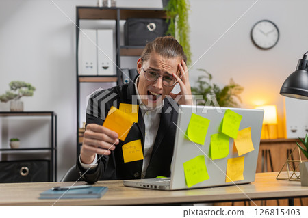 Tired exhausted businessman with laptop sticky notes with many tasks sitting at home office desk Tired exhausted businessman with laptop sticky notes with many tasks sitting at home office desk 126815403