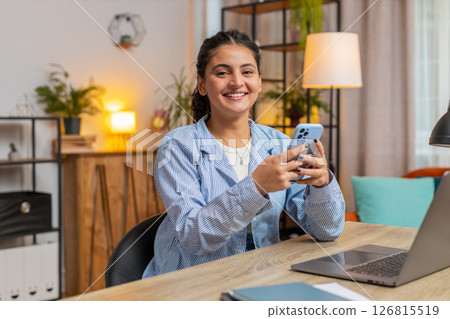 Smiling young woman freelancer sitting at table using smartphone texting in living room at home Smiling young woman freelancer sitting at table using smartphone texting in living room at home 126815519