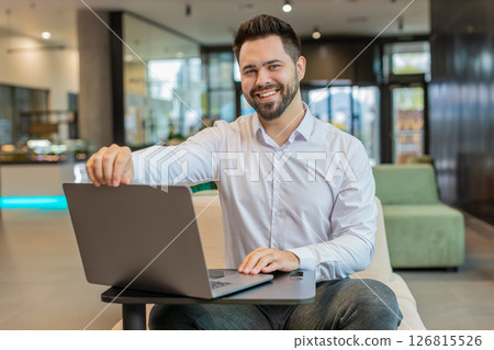 Young Caucasian businessman in while shirt opens laptop and starts working sitting in office lobby Young Caucasian businessman in while shirt opens laptop and starts working sitting in office lobby 126815526