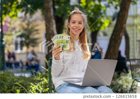 Happy rich woman using laptop winner victory holding cash money in euro banknotes sitting on chair Happy rich woman using laptop winner victory holding cash money in euro banknotes sitting on chair 126815658
