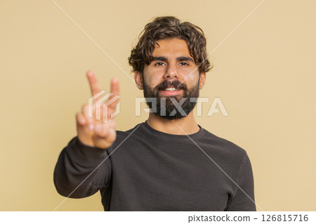 Young Indian man showing victory sign, doing peace gesture standing against beige studio background 126815716
