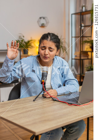 Young positive Indian woman blogger singer using laptop while singing into microphone at table 126815731
