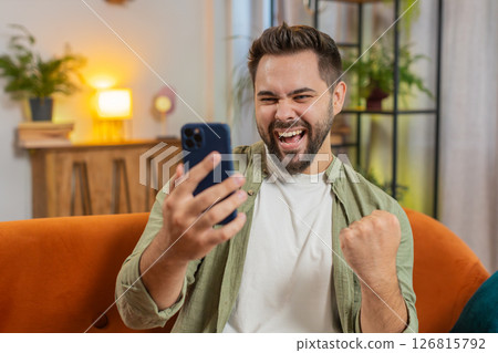 Happy young man using cellphone celebrating success victory winning lottery on sofa in living room Happy young man using cellphone celebrating success victory winning lottery on sofa in living room 126815792