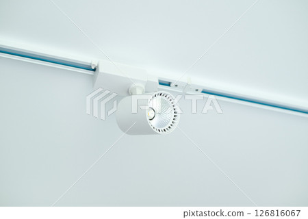 Modern white LED track light fixture mounted on ceiling rail, minimalistic interior lighting solution, close-up view. Modern white LED track light fixture mounted on ceiling rail, minimalistic interior lighting solution, close-up view. 126816067