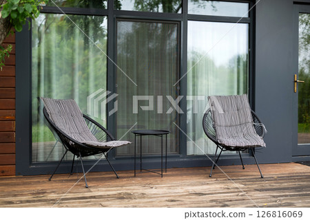 Two modern chairs and a small table on a wooden terrace by glass doors. Minimalistic outdoor seating area for relaxation. 126816069