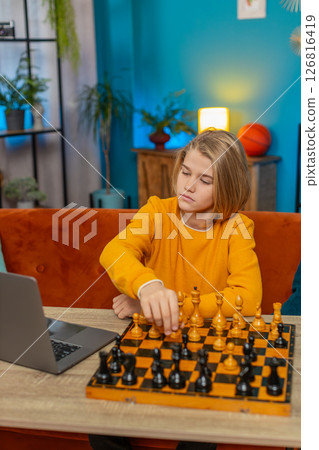 Teenager boy playing online chess on laptop with real chessboard thoughtfully moving pieces at table Teenager boy playing online chess on laptop with real chessboard thoughtfully moving pieces at table 126816419