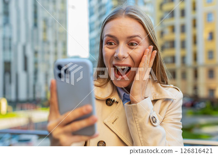 Caucasian mature woman looking shocked while reading good news on smartphone on downtown city street Caucasian mature woman looking shocked while reading good news on smartphone on downtown city street 126816421
