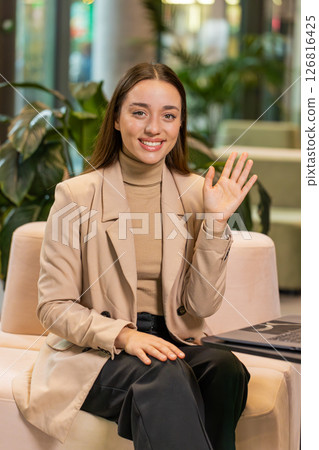 Portrait of smiling young businesswoman with laptop wave hand at camera sitting in hotel lobby Portrait of smiling young businesswoman with laptop wave hand at camera sitting in hotel lobby 126816425
