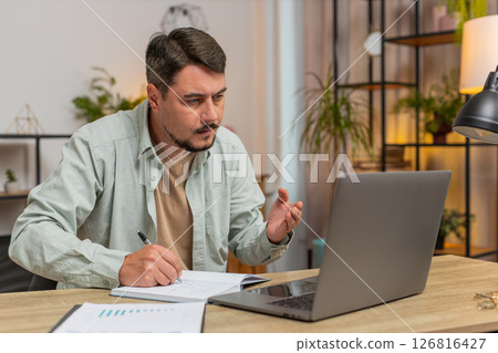 Young man writing down notes while attending online office meeting on laptop at home office desk Young man writing down notes while attending online office meeting on laptop at home office desk 126816427