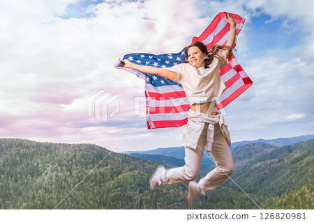 A young woman exuberantly jumps in the air, expressing her patriotic spirit as she waves the American flag. Dressed in casual white attire, she stands on a scenic overlook surrounded by lush greenery A young woman exuberantly jumps in the air, expressing her patriotic spirit as she waves the American flag. Dressed in casual white attire, she stands on a scenic overlook surrounded by lush greenery 126820981