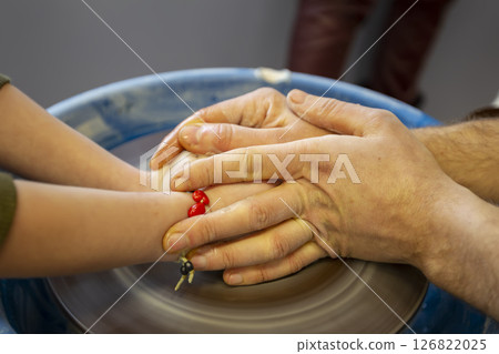 Close-up of a potter's hands and a child's hand with an item on a potter's wheel. Working with clay. Clay workshop. Craft training. Close-up of a potter's hands and a child's hand with an item on a potter's wheel. Working with clay. Clay workshop. Craft training. 126822025