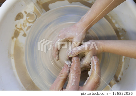 Close-up of a potter's hands and a child's hand with an item on a potter's wheel. Working with clay. Clay workshop. Craft training. Close-up of a potter's hands and a child's hand with an item on a potter's wheel. Working with clay. Clay workshop. Craft training. 126822026