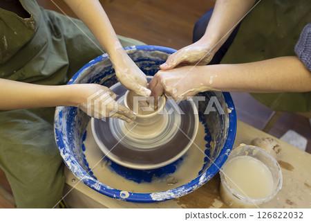 Close-up of a potter's hands and a child's hand with an item on a potter's wheel. Working with clay. Clay workshop. Craft training. 126822032
