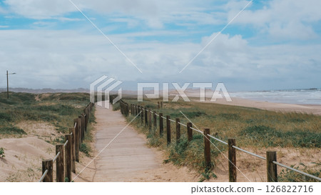 Wooden boardwalk path leading to a serene sandy beach with grassy dunes on both sides Wooden boardwalk path leading to a serene sandy beach with grassy dunes on both sides 126822716