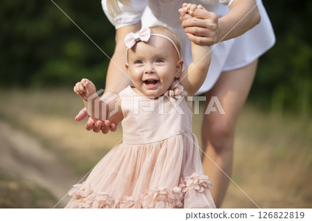 A happy one-year-old girl is holding her mother's hands. Little child for a walk. Sweet baby learns to walk. 126822819