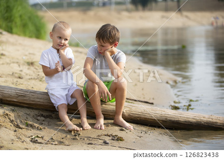 Two brothers, the elder and the younger, are playing by the river. Children on the sandy shore talk and indulge. 126823438