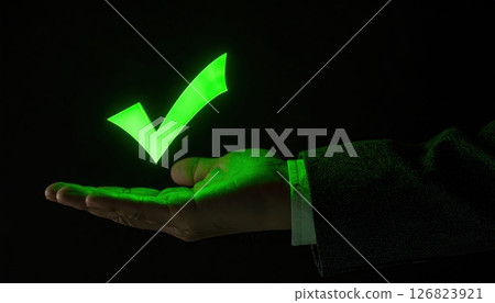 Hand holding a glowing green checkmark symbol highlighting approval or success in dark background 126823921