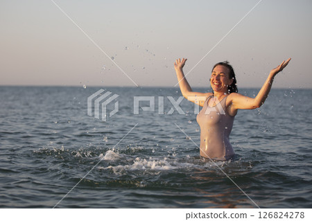 Happy woman at the sea in the sun. Woman in a swimsuit splashes water on a background of waves. Summer vacation on the beach. 126824278