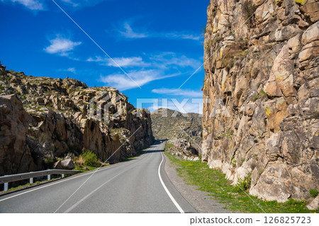 Asphalt road passing through stone cliffs at Stone Gates Chuysky Trakt Altai Russia. Iconic mountain pass framed by towering rocky formations. Asphalt road passing through stone cliffs at Stone Gates Chuysky Trakt Altai Russia. Iconic mountain pass framed by towering rocky formations. 126825723