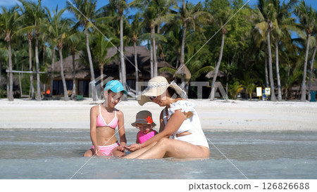 Mom sits in ocean water with daughters on sunny tropical beach enjoying summer day 126826688