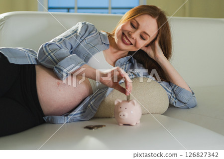 Pregnancy motherhood saving money for future investment. Pregnant woman with big belly putting coin into piggy bank at home. Pregnancy girl with piggy bank. Maternity benefit family planning concept Pregnancy motherhood saving money for future investment. Pregnant woman with big belly putting coin into piggy bank at home. Pregnancy girl with piggy bank. Maternity benefit family planning concept 126827342