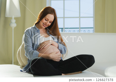 Pregnancy motherhood people expectation future. Pregnant woman touching big belly sitting on couch at home. Girl hugging her tummy enjoying pregnancy. Maternity tenderness parenthood new life concept Pregnancy motherhood people expectation future. Pregnant woman touching big belly sitting on couch at home. Girl hugging her tummy enjoying pregnancy. Maternity tenderness parenthood new life concept 126827378