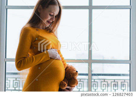Pregnancy motherhood. Pregnant woman with big belly hugging teddy bear standing near window at home. Happy smiling girl enjoying pregnancy. Maternity tenderness parenthood new life concept 126827403