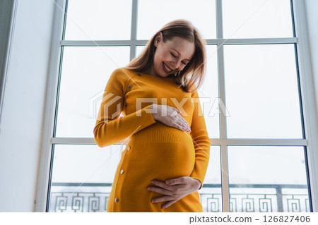 Pregnancy motherhood people expectation future. Pregnant woman with big belly standing near window at home. Girl hugging her tummy enjoying pregnancy. Maternity tenderness parenthood new life concept 126827406