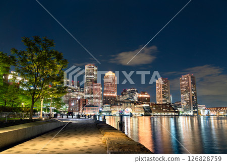 View of downtown Boston illuminated at night as seen from Fan Pier Park, with reflections shimmering across the calm harbor water and a tree-lined promenade in the foreground. 126828759