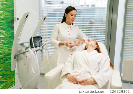 Cosmetologist in white uniform consulting with woman patient lying on bed by window, discussing various beauty treatments in modern beauty clinic. Concept of innovative healthcare technology. Cosmetologist in white uniform consulting with woman patient lying on bed by window, discussing various beauty treatments in modern beauty clinic. Concept of innovative healthcare technology. 126829361