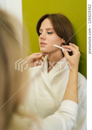 Vertical shot of professional beautician marking client facial contours with white pencil, preparing permanent makeup procedure with symmetry and precision in modern beauty salon. Vertical shot of professional beautician marking client facial contours with white pencil, preparing permanent makeup procedure with symmetry and precision in modern beauty salon. 126829362