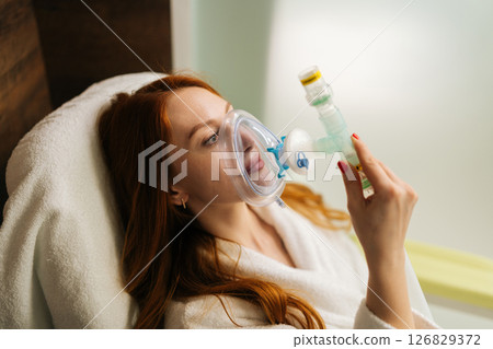 Close-up face of sick redhead female patient wearing oxygen mask connected to tube, breathing with difficulties, during respiratory therapy in hospital. Procedure increases body adaptation abilities. 126829372